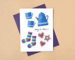 Load image into Gallery viewer, Greeting card with blue teacups, socks, and decorative elements on a beige background with text 'cosy on down'

