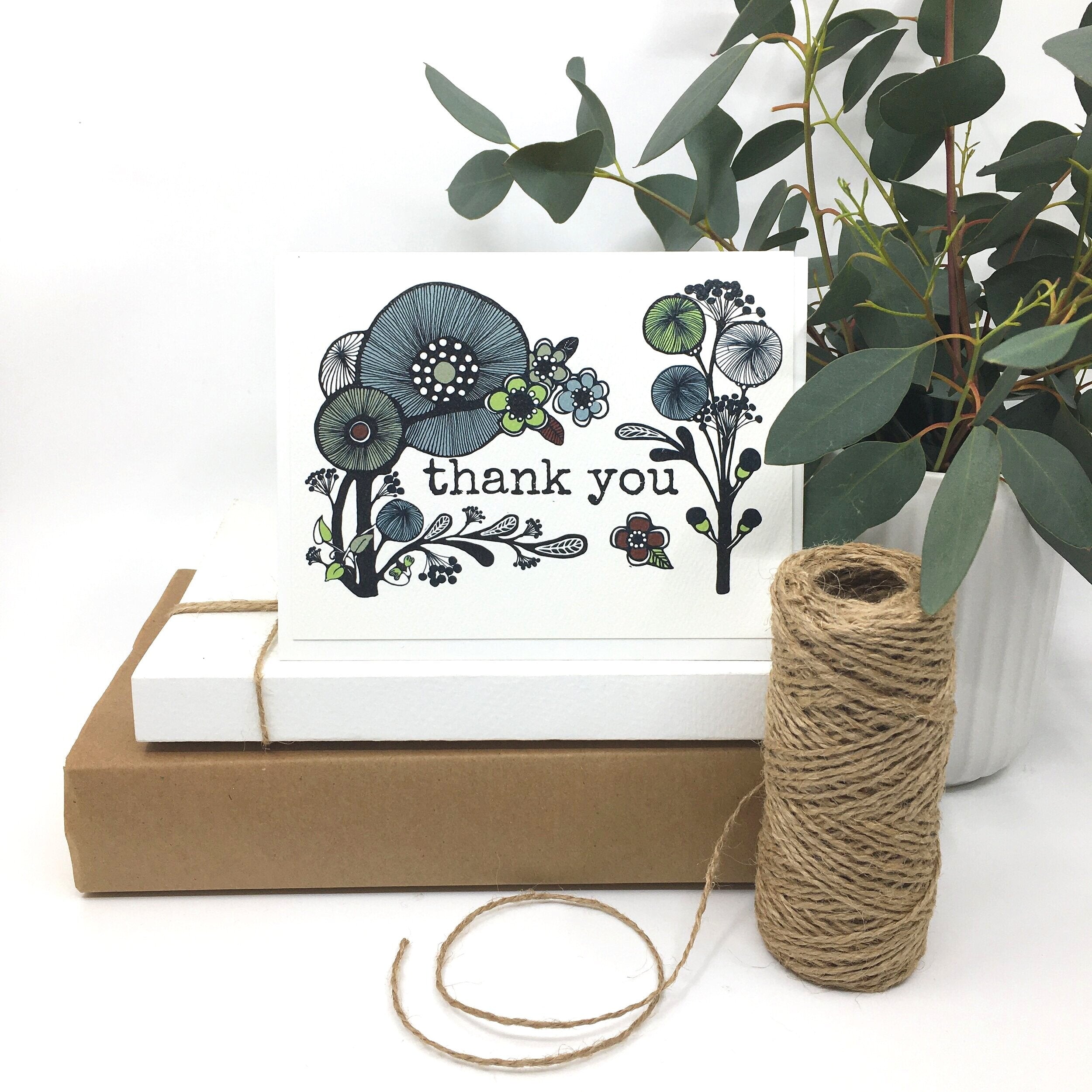 Scandinavian folk style thank you card sits on two paper boxes besides a roll of twine and a potted plant