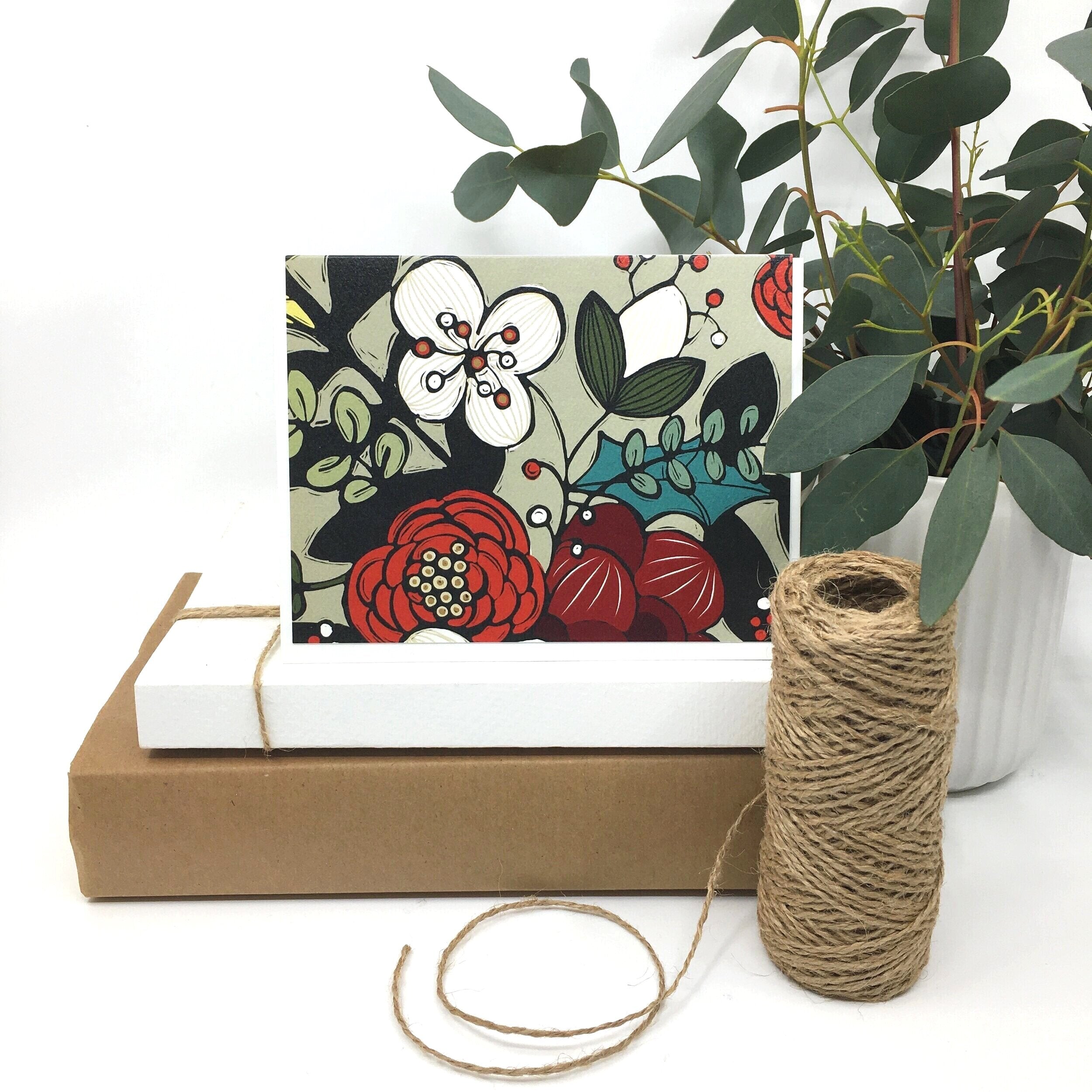 red and white floral card sits on two paper boxes besides a roll of twine and a potted plant