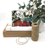 Load image into Gallery viewer, red and white floral card sits on two paper boxes besides a roll of twine and a potted plant
