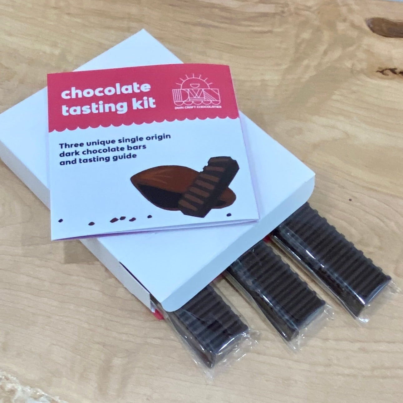 three chocolate bars pop out of a white box with the chocolate tasting guidebook resting on the box top on a wooden background
