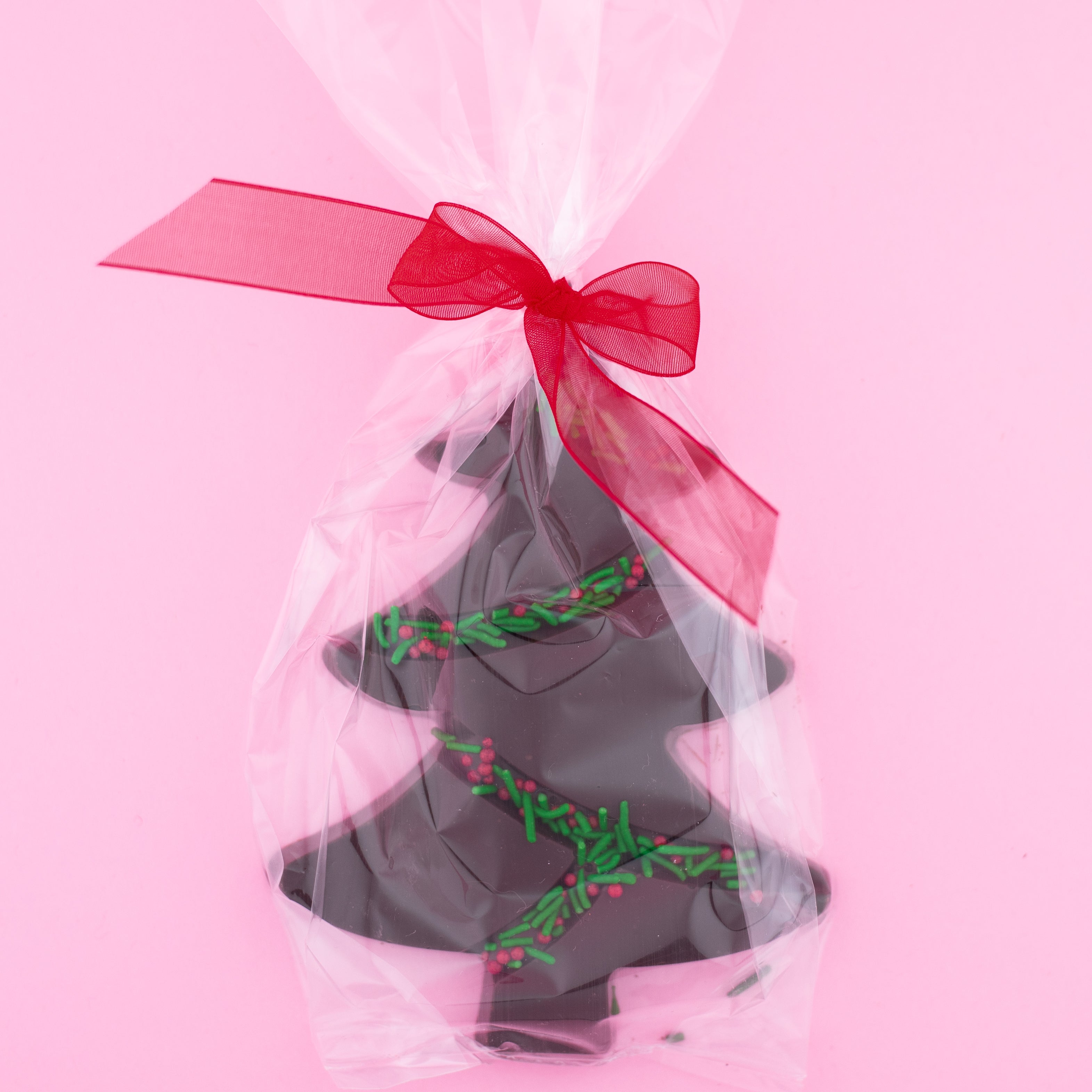 dark chocolate tree with green and red sprinkles in a clear bag with a red bow on a pink background