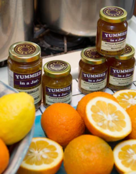 Jars of lemon and ginger marmalade with the label 'Yummies in a Jar' on a kitchen countertop, surrounded by sliced oranges and a whole lemon.
