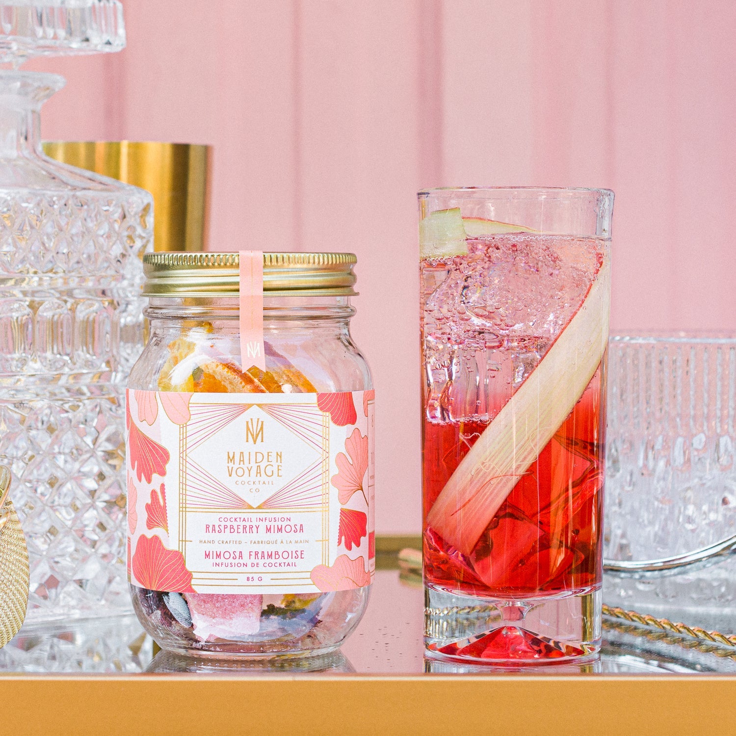 raspberry mimosa cocktail infusion kit jar next to a tall glass full with the cocktail, ice and a slice of sugar cane