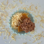 Load image into Gallery viewer, a single coconut cluster leans against a piece of toffee and a piles of toasted coconut in a blue bowl
