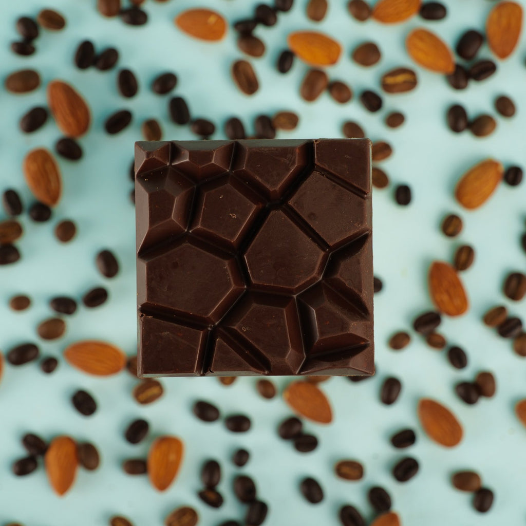 a mocha almond crunch bar lays on a bed of almonds and coffee beans on a blue background